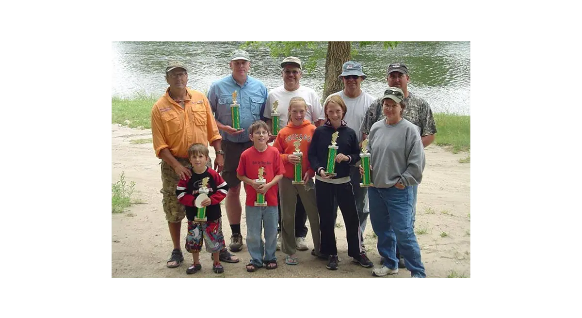 Group of adults and children standing in front of water holding trophies, the first kayak fishing tournament winners